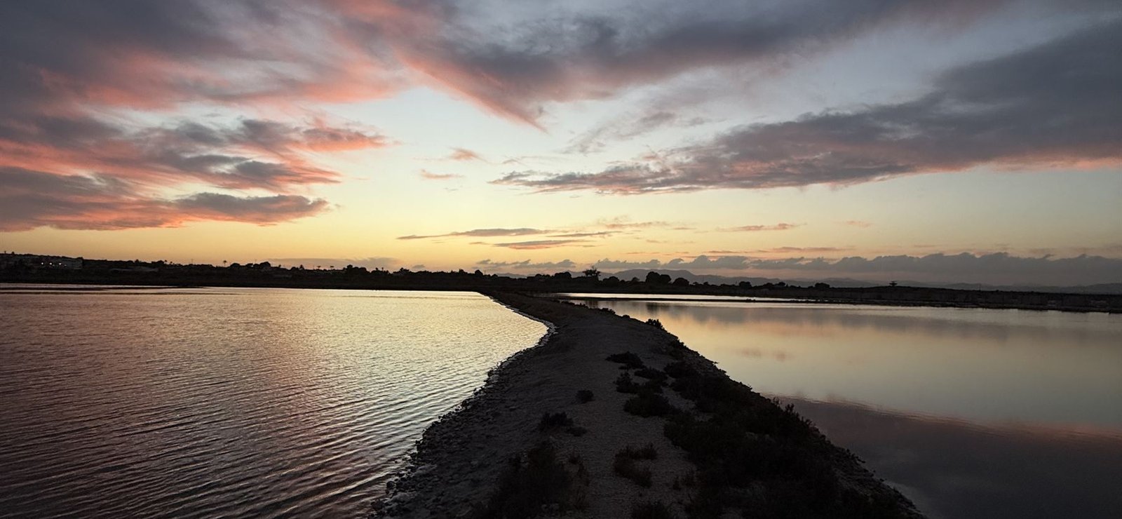 Dique de unas salinas al atardecer, un camino recto entre dos lagunas hacia el horizonte.